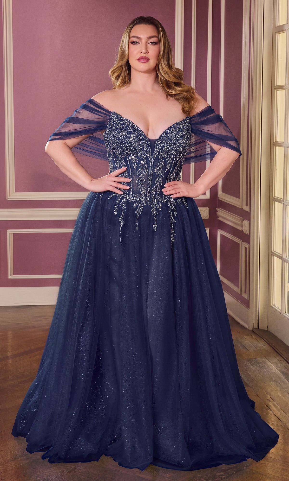 A woman stands in a room with pink walls and white trim, wearing the Plus-Size Glitter Prom Ball Gown (CLCD721C) in navy. The off-the-shoulder, floor-length dress features sparkling beaded embellishments.